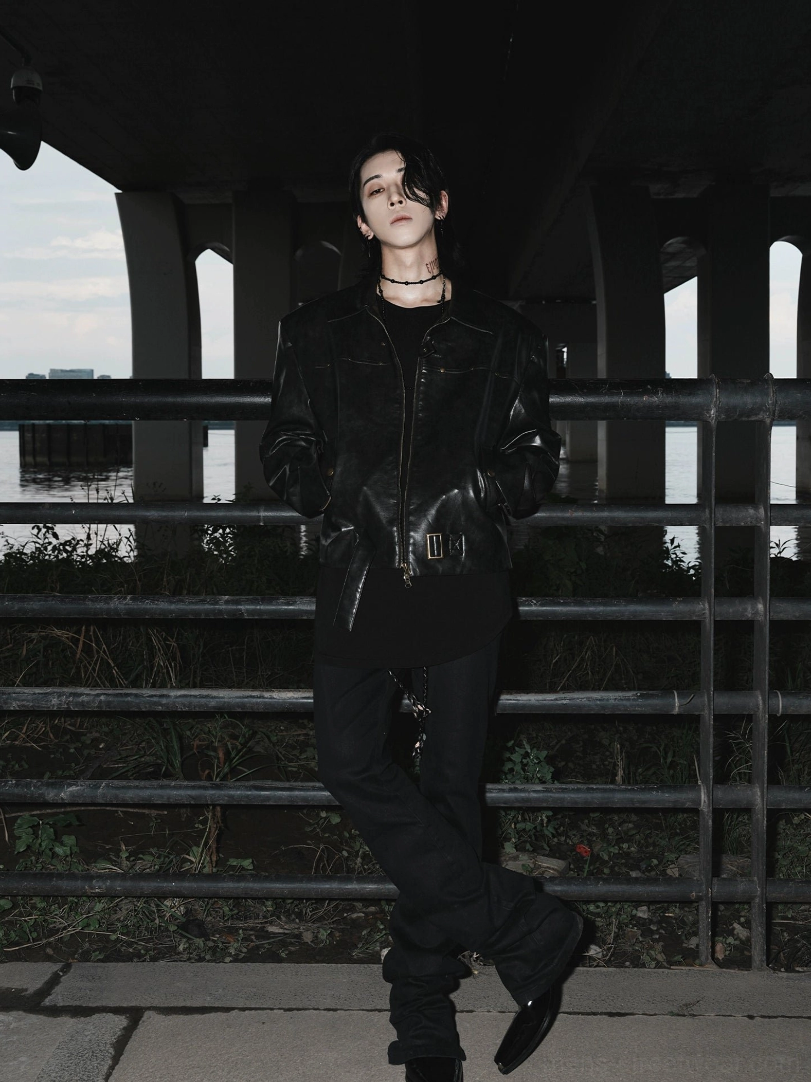 Full-body model wearing a black oversized faux leather moto jacket with all-black streetwear outfit in an urban under-bridge setting