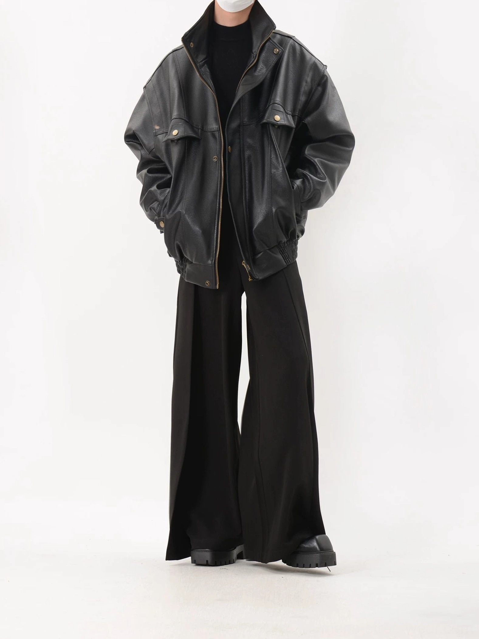 Full-body front styling of black oversized faux leather utility bomber jacket with high collar, brass-tone snap buttons, chest flap pockets, side pockets, and wide-leg pants