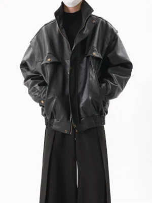 Three-quarter front view of a black oversized faux leather utility bomber jacket with dropped shoulders, high stand collar, flap chest pockets, antique-tone snap buttons, elasticated hem, and matte streetwear finish