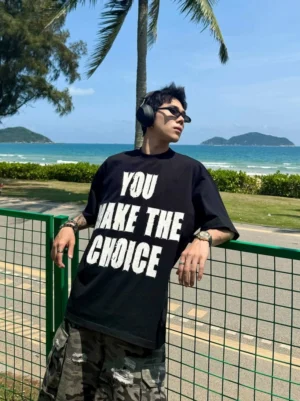 Side view of loose fit black graphic T-shirt featuring bold white choice slogan, worn with ripped camouflage pants beside a coastal highway