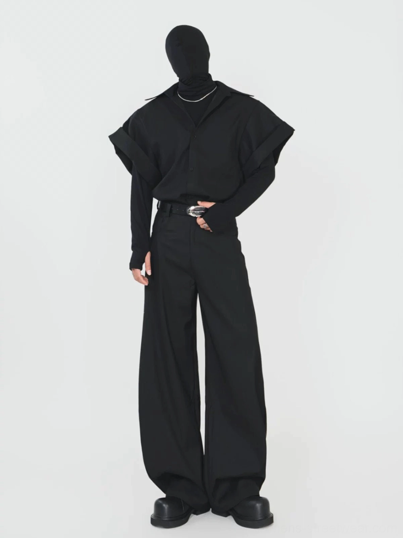 Full body view of a man wearing a black oversized layered shirt jacket with wide short sleeves, wide leg trousers, and silver buckle belt
