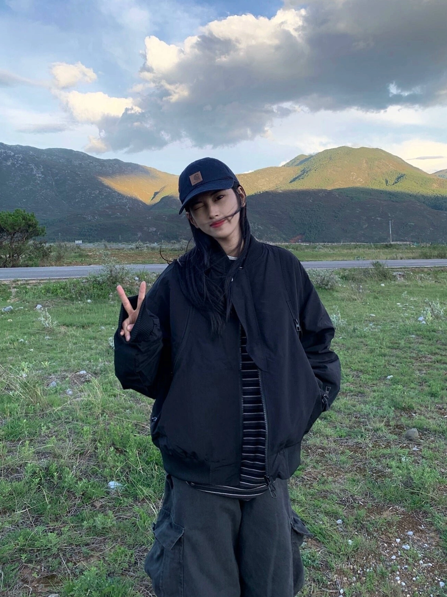 Black oversized matte flight bomber jacket front view styled with striped shirt at sunset mountain roadside