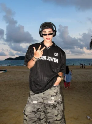 Front view of a male model wearing a black oversized mesh football jersey tee with vertical stripes, distressed 08 graphic, contrast polo collar, sunglasses, and headphones on a coastal beach