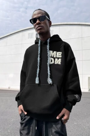 Black oversized unisex hoodie with raised white puff print lettering, frayed woven drawstrings, kangaroo pocket, and streetwear styling on a model