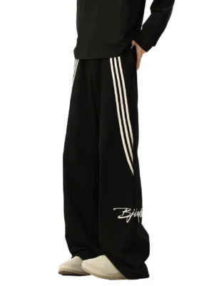 Black oversized wide-leg track pants with contrasting white side stripes, white script embroidery, relaxed Korean streetwear silhouette, and casual athletic styling