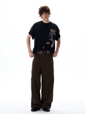 Black oversized graphic T-shirt with retro passport stamps and travel patch prints styled with brown wide-leg denim