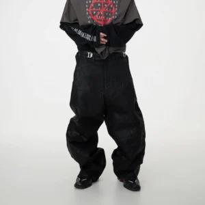 Front three-quarter view of black oversized patchwork baggy jeans with coated denim, geometric panel seams, wide legs, stacked ankles, and utility buckle details styled with layered streetwear