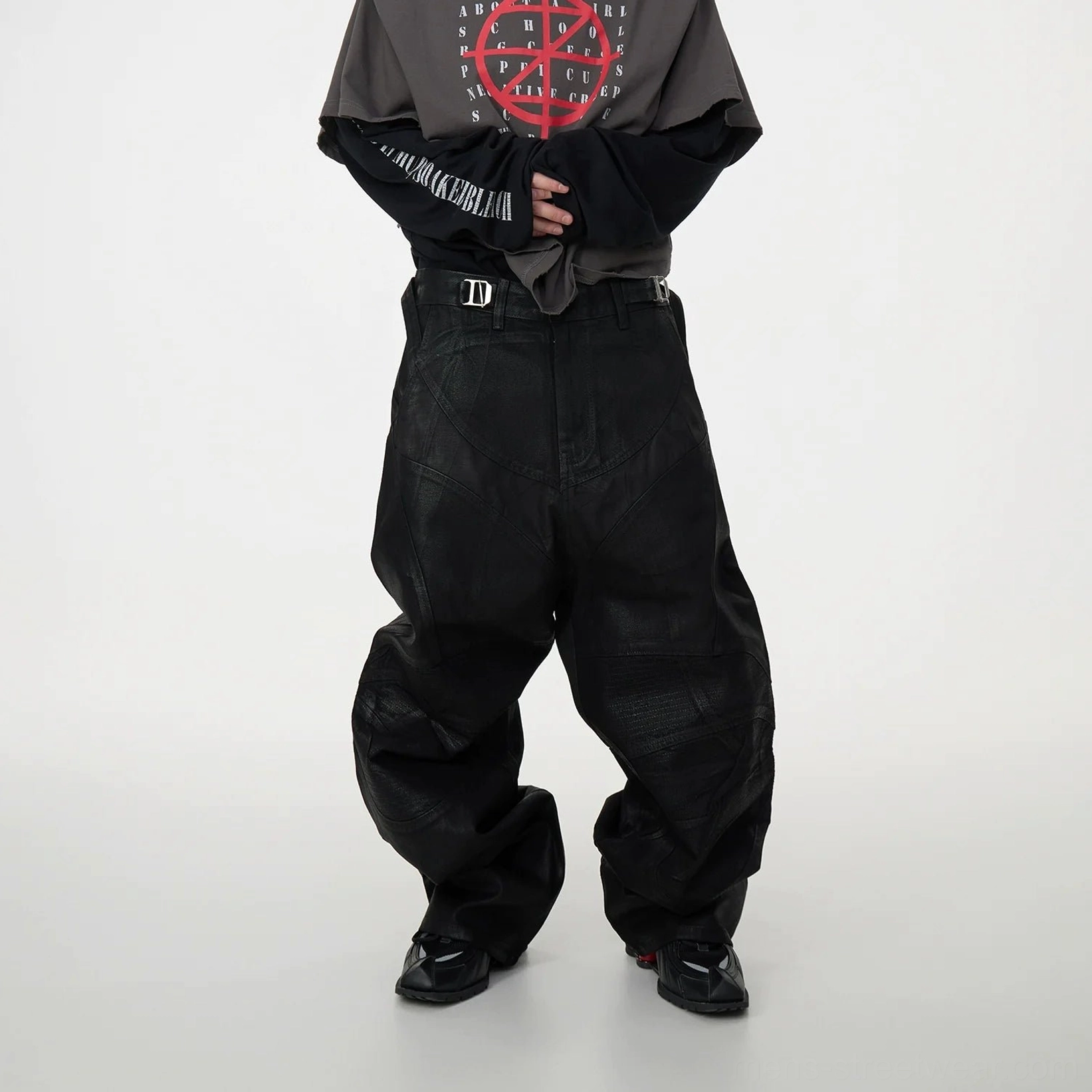 Front three-quarter view of black oversized patchwork baggy jeans with coated denim, geometric panel seams, wide legs, stacked ankles, and utility buckle details styled with layered streetwear
