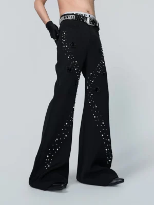 Three-quarter view of black pearl-flare trousers showing flowing rhinestone-inspired leg trims, raised floral appliques, high waist, and tailored streetwear silhouette