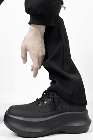 Black platform casual sneakers shown in side profile with black trousers, highlighting the oversized chunky rubber sole and matching black laces