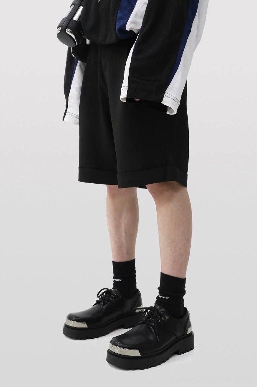 Male model wearing black platform derby shoes with silver toe caps, black socks, rolling shorts, and a color-block streetwear jacket