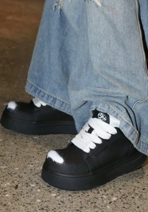 Black platform derby shoes worn with loose blue jeans on a concrete floor, featuring white contrast laces and spray-finish toe details