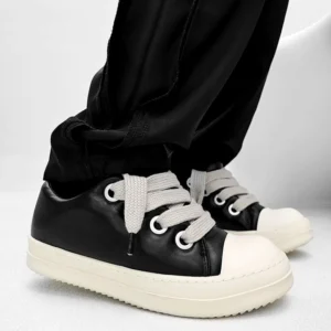 Black platform sneakers worn with loose black trousers, featuring a smooth leather upper, off-white rounded toe cap, chunky cream sole, and wide flat laces