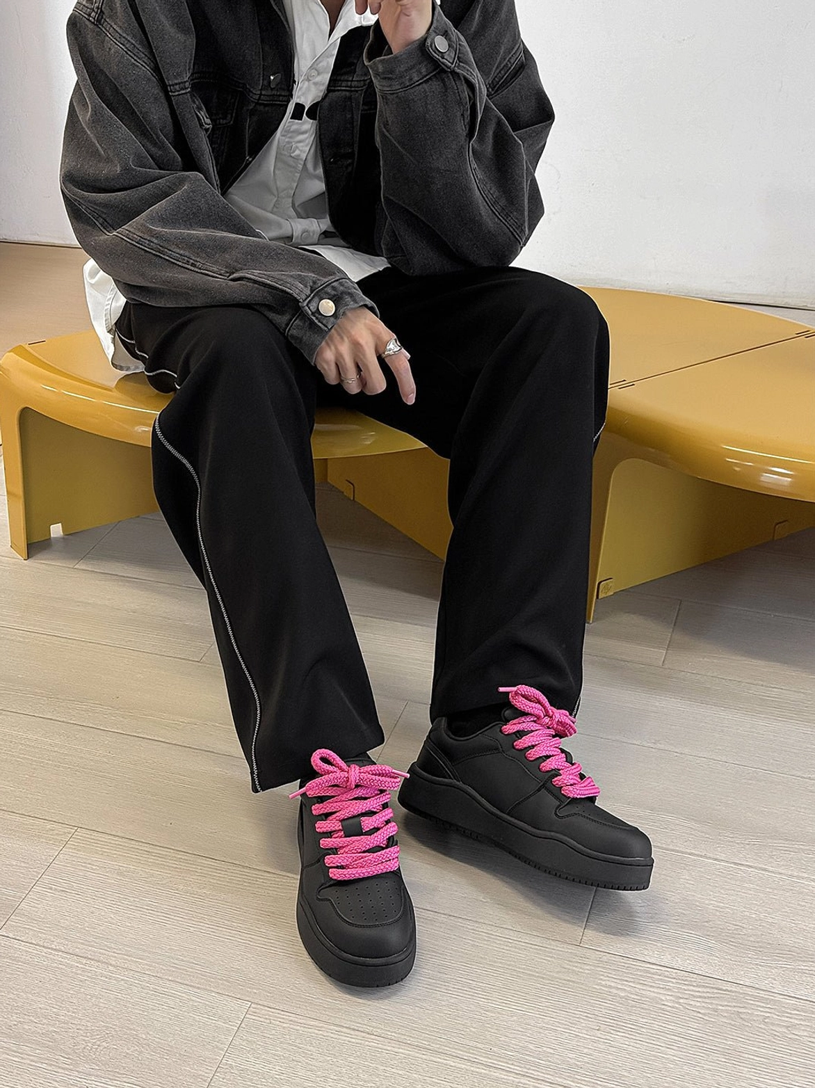 Seated male model wearing black platform sneakers with vivid hot pink laces denim jacket and wide leg pants