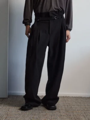 Close front view of black pleated belted wide-leg trousers with high waist, metal belt hardware, and relaxed tailored drape