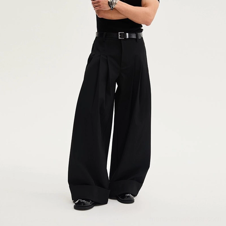 Black pleated wide-leg dress trousers shown from a low front angle with tailored pleats, rolled cuffs, belt styling, and polished formal shoes