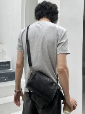 Black PU leather barrel crossbody satchel worn over shoulder by male model in minimalist stairwell rear view