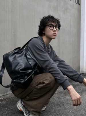 Black PU leather sling shoulder bag worn on the back with a relaxed oversized streetwear outfit against a concrete urban wall