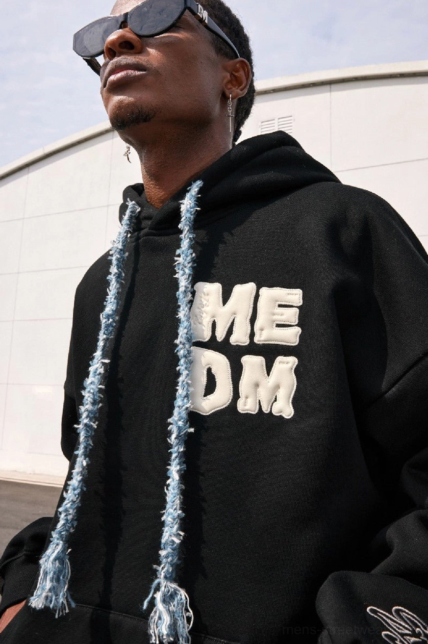 Close-up of black oversized streetwear hoodie showing dimensional raised chest lettering, textured drawstrings, and subtle sleeve embroidery