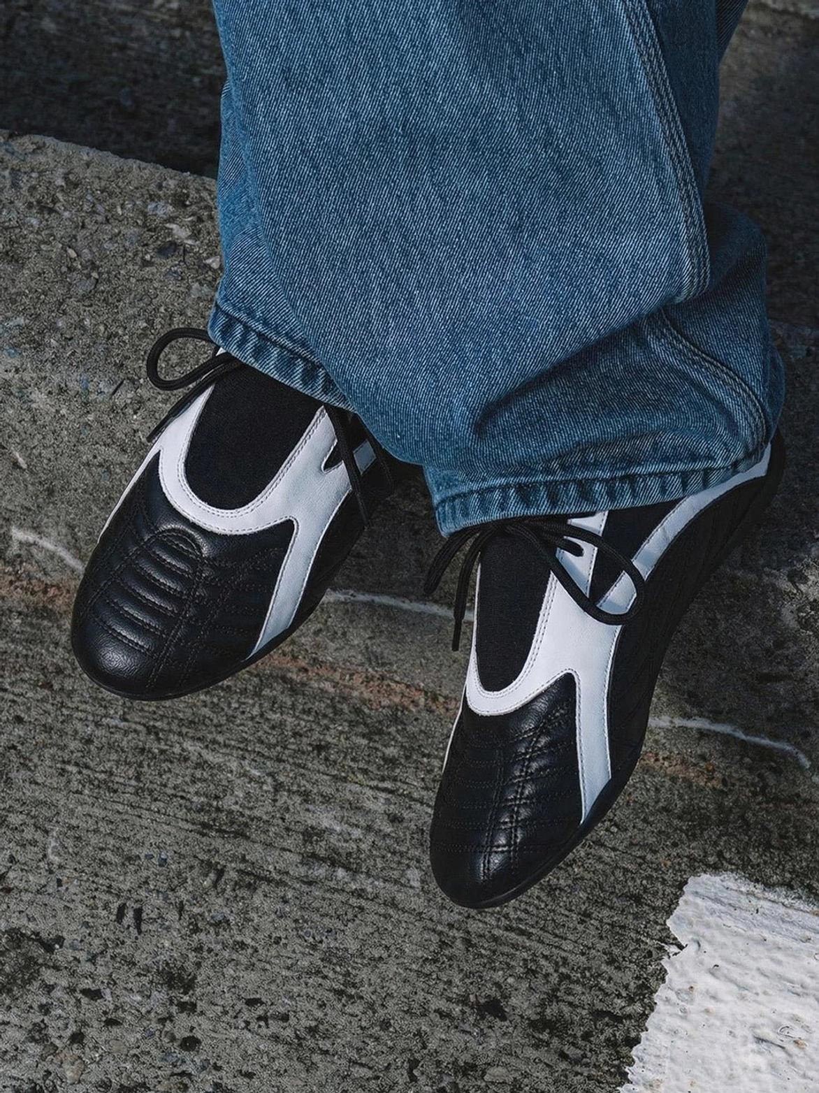 Black quilted panel flat sneakers styled with blue denim jeans, white contrast side panels, black laces, and a split rubber sole