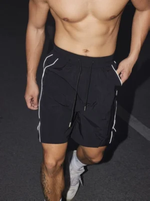 Black reflective stripe streetwear swim shorts with drawstring elastic waist photographed from a low front angle on an urban night street
