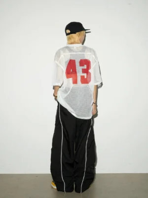 Back view of a model wearing black reflective wide-leg cargo pants with white side piping, a graphic mesh streetwear top, baseball cap, and sneakers in a studio setting