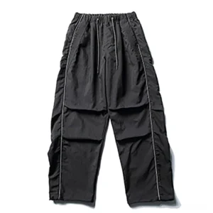 Front flat lay of black reflective utility cargo pants showing elastic drawstring waistband, pleated panels, smooth woven fabric, white reflective side piping, and loose wide-leg cut