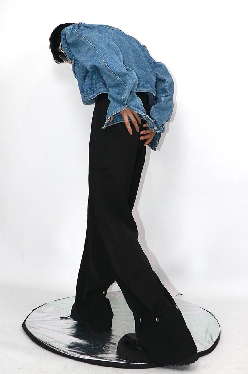 Back view of black relaxed straight-leg trousers with standard waistband, metal snap hem details, cropped denim jacket, and platform shoes