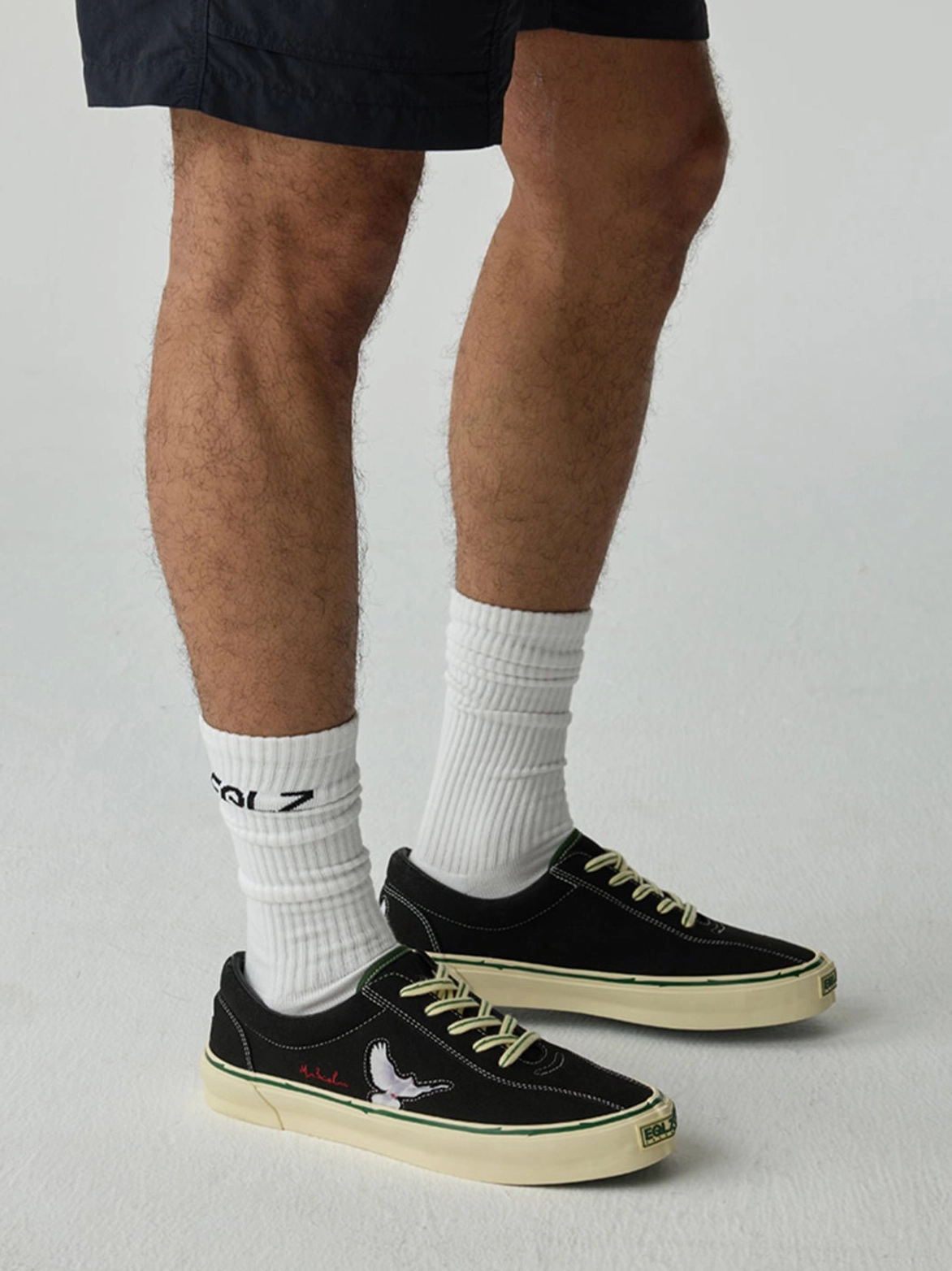 Male model wearing black rose embroidered skate shoes with black shorts and white crew socks in a streetwear styling shot