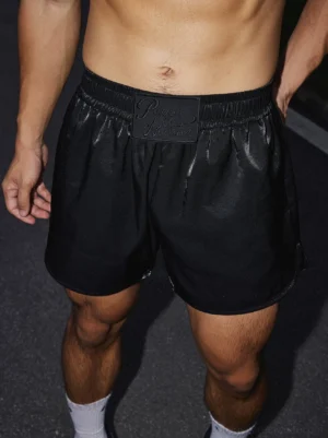 Low-angle close-up of black satin logo athletic shorts with elastic waistband and relaxed streetwear fit