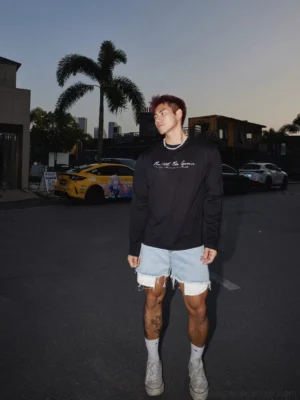 Full-body model wearing a black script print long sleeve streetwear tee with ripped denim shorts, white socks, and casual canvas sneakers in an urban dusk setting