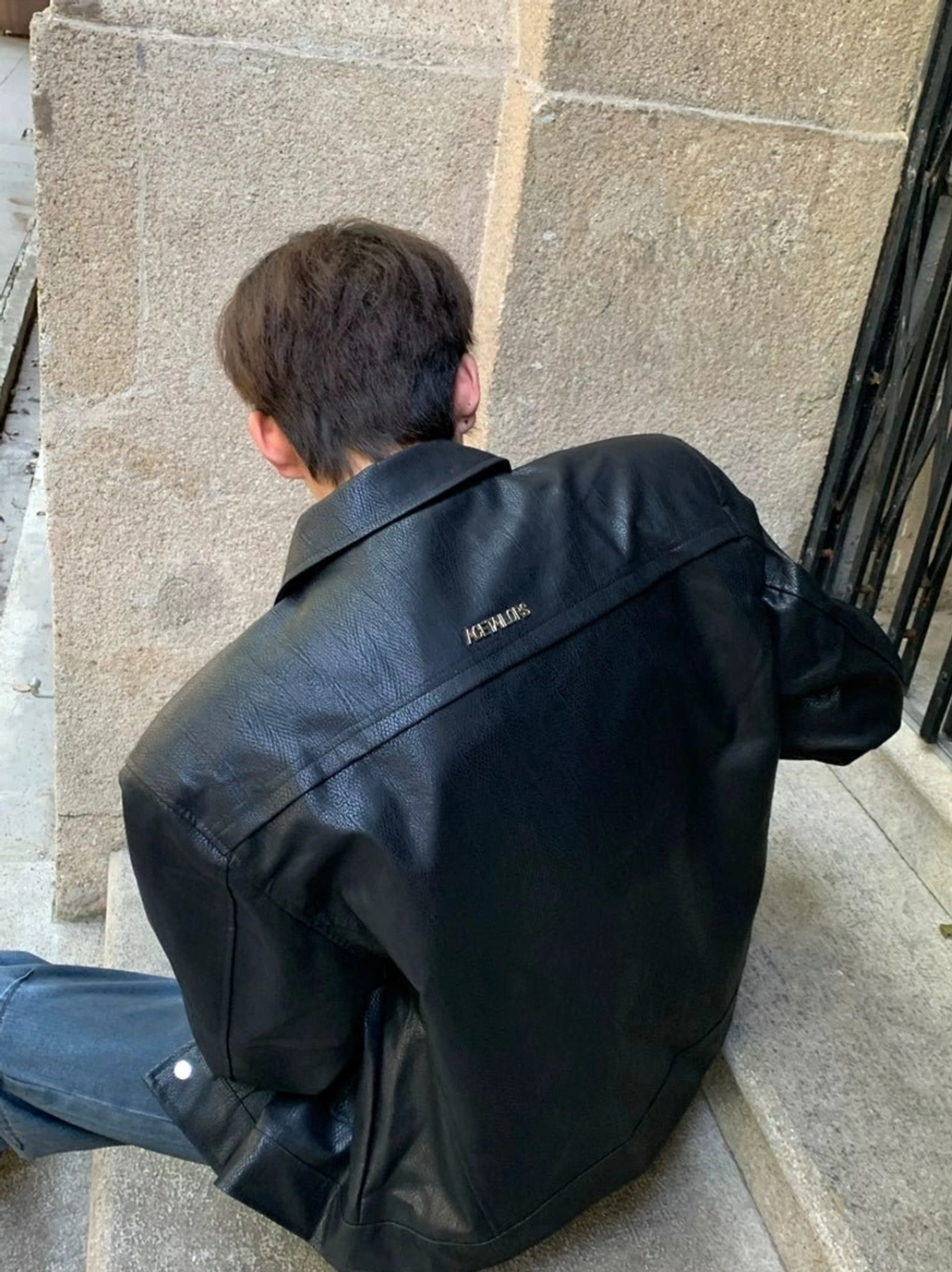Black sculpted wide-shoulder jacket shown from the back on a model seated on stone steps in an urban streetwear setting