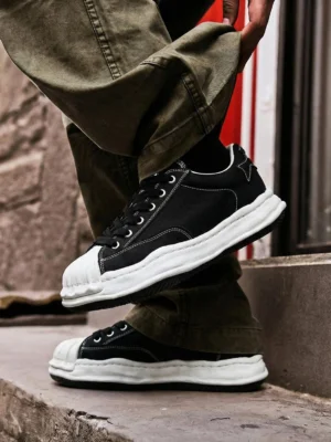 Black shell-toe court sneakers worn with olive cargo pants on outdoor stairs showing the thick white court sole