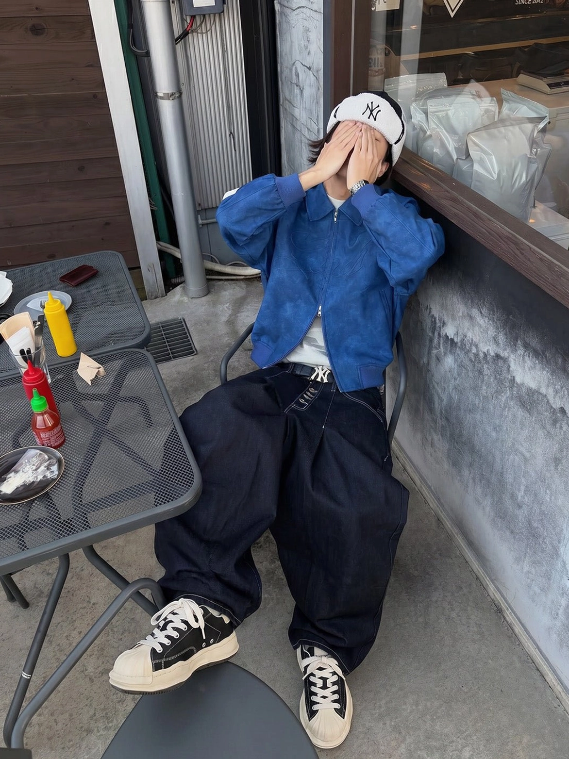 Male model wearing black shell-toe platform sneakers with baggy dark jeans and a blue jacket in a casual Seoul-inspired streetwear setting