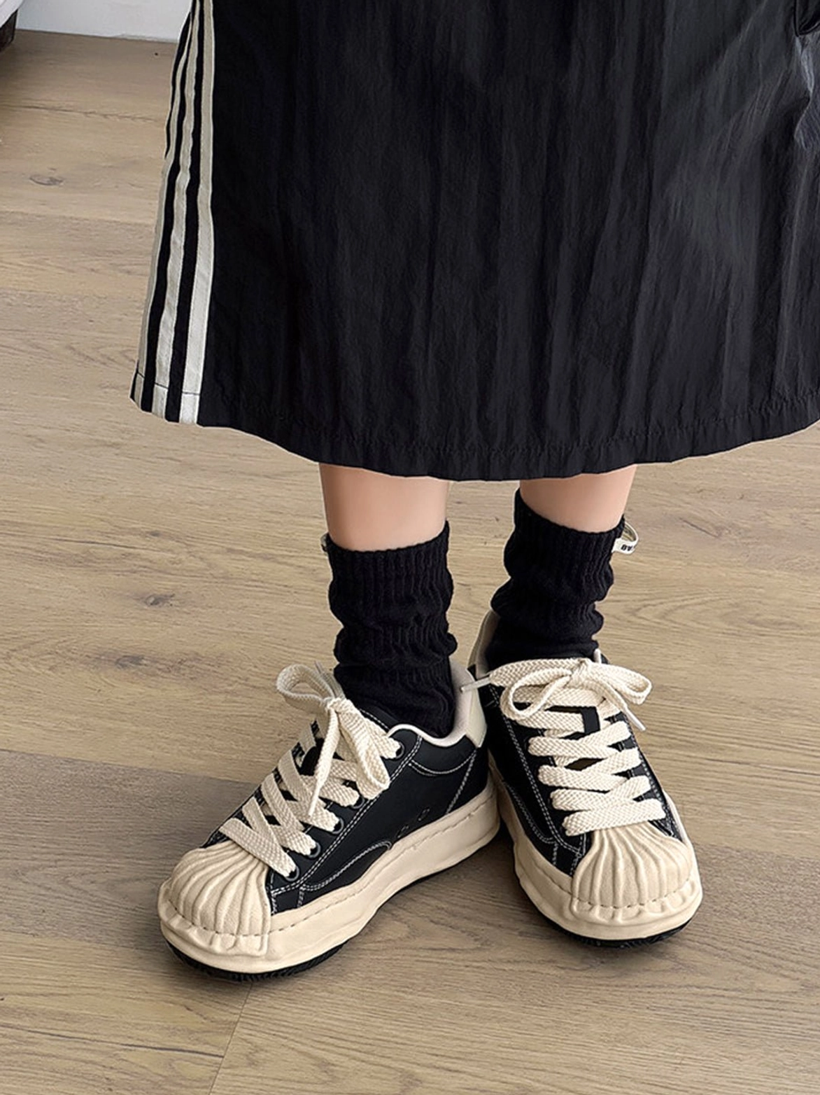 Female model wearing black shell-toe platform sneakers with black slouch socks and midi skirt