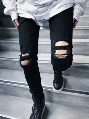 Close-up front view of black shredded knee skinny jeans with layered frayed rips, stacked hems, black platform sneakers, and oversized white streetwear top