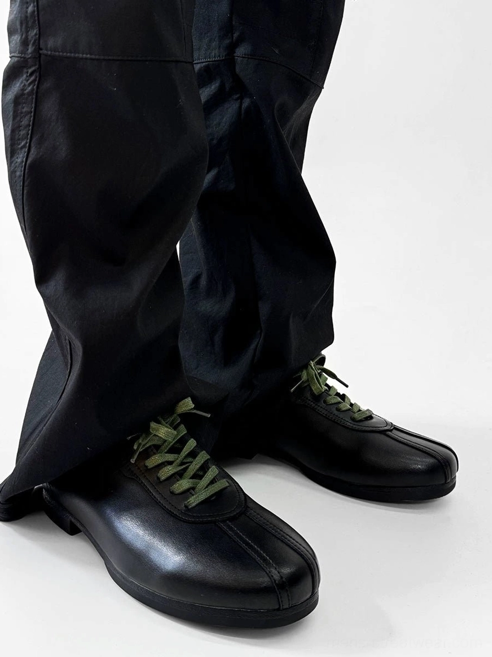 Three-quarter view of black split-toe derby shoes showing split toe stitching, green flat laces, and low block heel