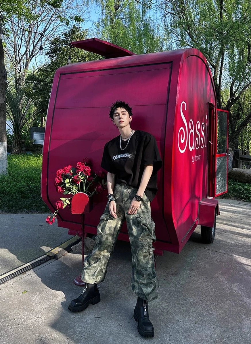 Male streetwear model wearing black square-toe front-zip platform boots with a black tee, camouflage cargo pants, and silver neck accessories