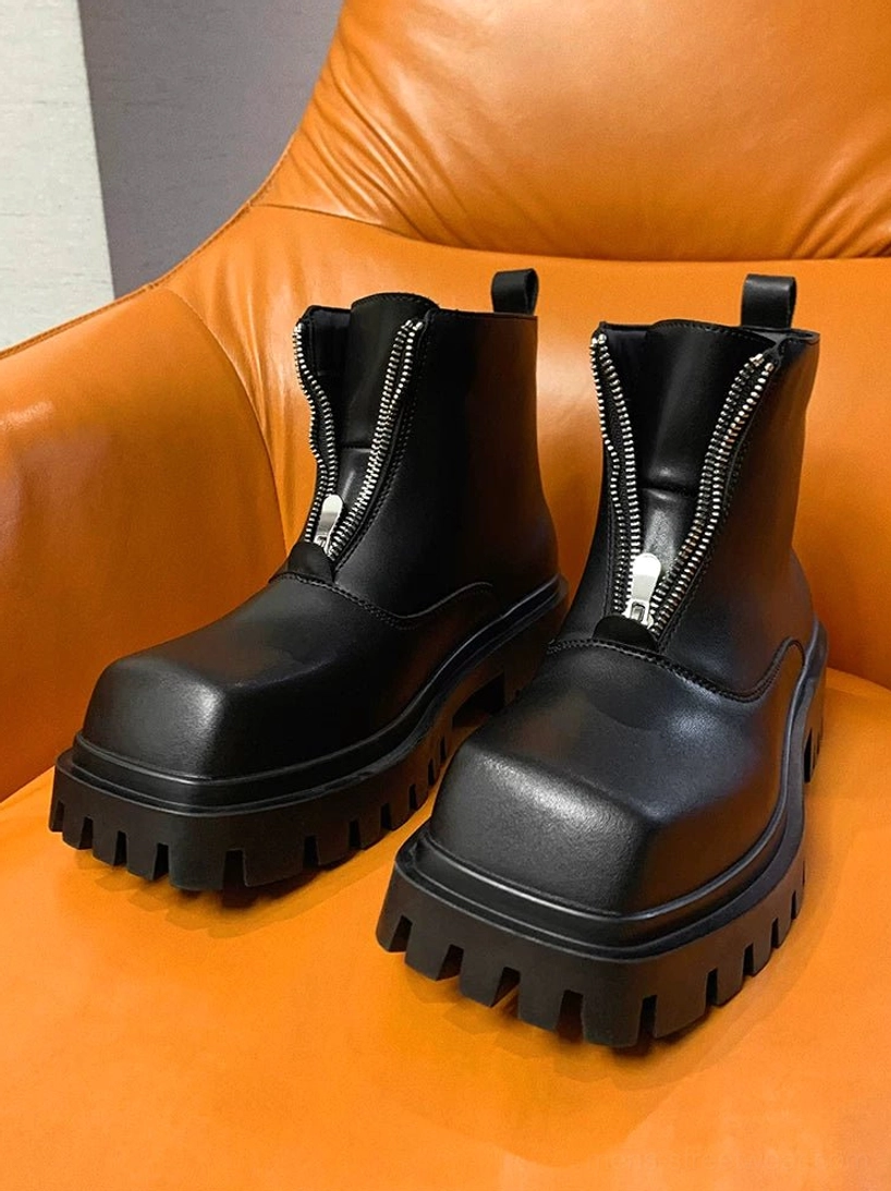 Black square-toe platform boots arranged on an orange leather chair, highlighting symmetrical stitching and the fully zipped front closure
