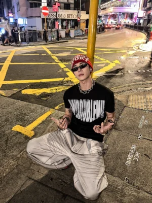 Black statement graphic streetwear T-shirt styled with loose gray trousers, red beanie, sunglasses, and urban Hong Kong night street fashion