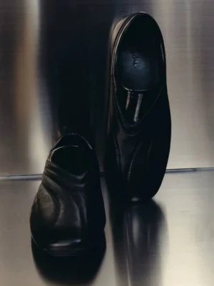 Pair of black low-profile leather slip-on shoes with curved wave stitching and glossy collar displayed inside a metal elevator
