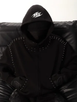 Black studded oversized full-zip hoodie close-up with silver metal shoulder studs, structured hood, graphic hood emblem, and tactical streetwear styling