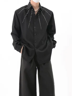 Black studded oversized streetwear overshirt shown from the front with radial silver-tone chest details, pointed collar, and relaxed long-sleeve silhouette