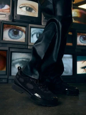 Model wearing black suede chunky low-top sneakers with black leather pants in a bold Seoul streetwear outfit against retro television monitors