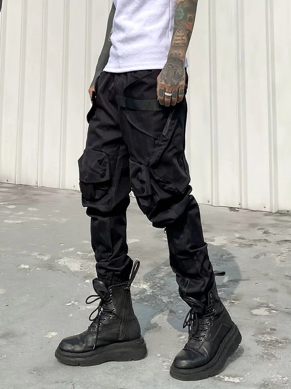 Close front view of black tactical cargo pants featuring large flap thigh pockets, slim waist strap detail, gathered cuffs, and platform work boots