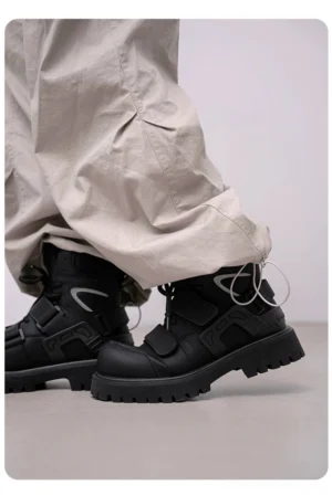 Black tactical work boots shown from a rear three-quarter angle with padded mid-calf shafts and loose beige cargo pants in a streetwear studio setting