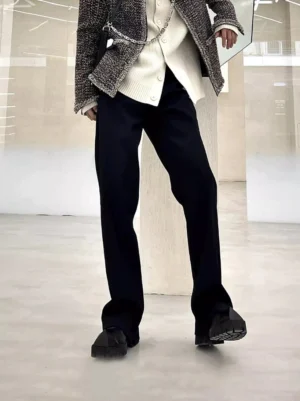 Men's black tailored straight-leg trousers in a low-angle front view, styled with a textured tweed coat and knit cardigan in a minimalist showroom