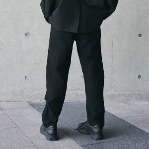 All-black technical track pants shown from the front with relaxed straight leg silhouette styled with black work boots