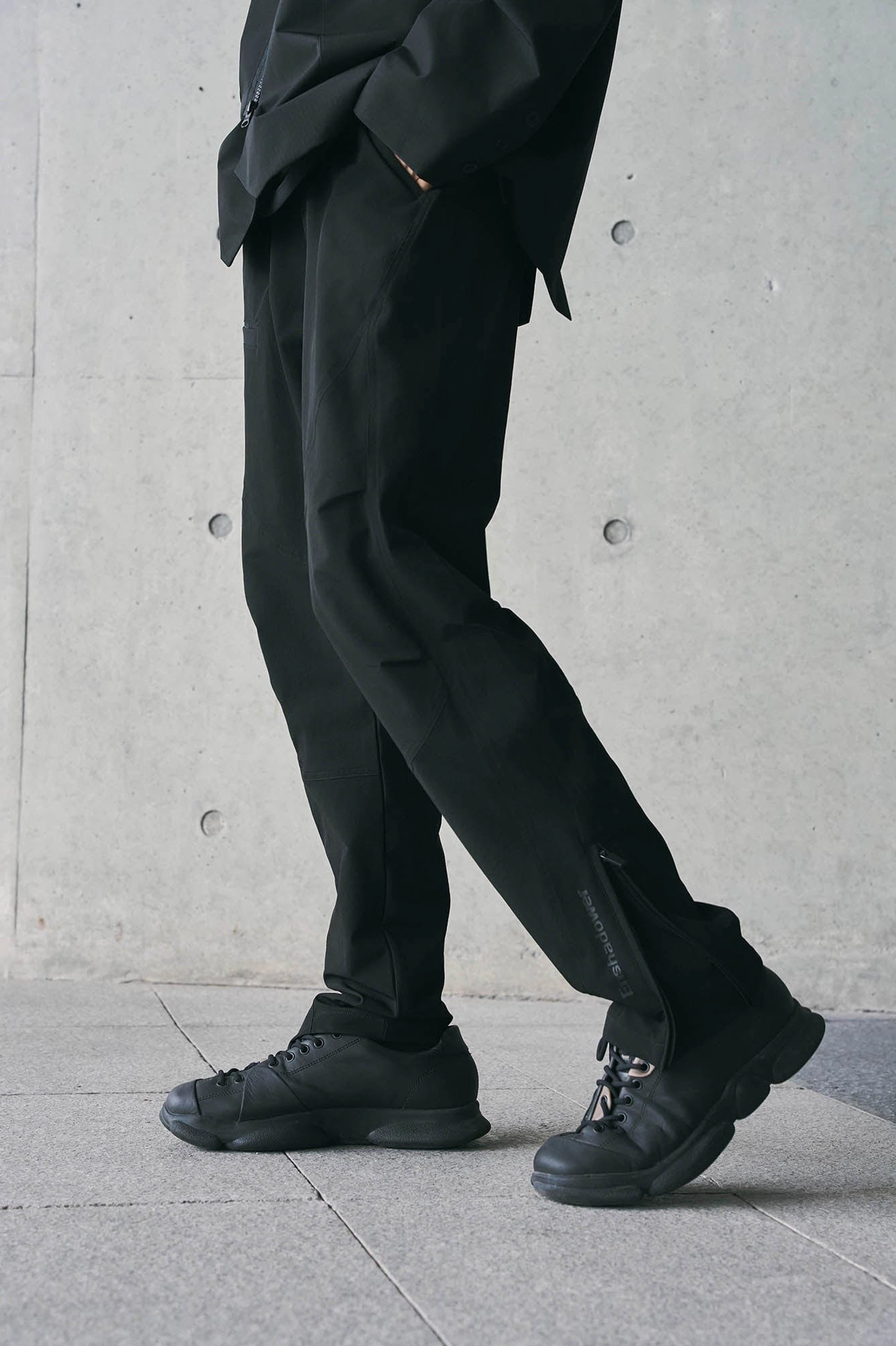 Side detail view of black technical track pants showing exposed ankle zipper closure and tapered hem with lace-up shoes