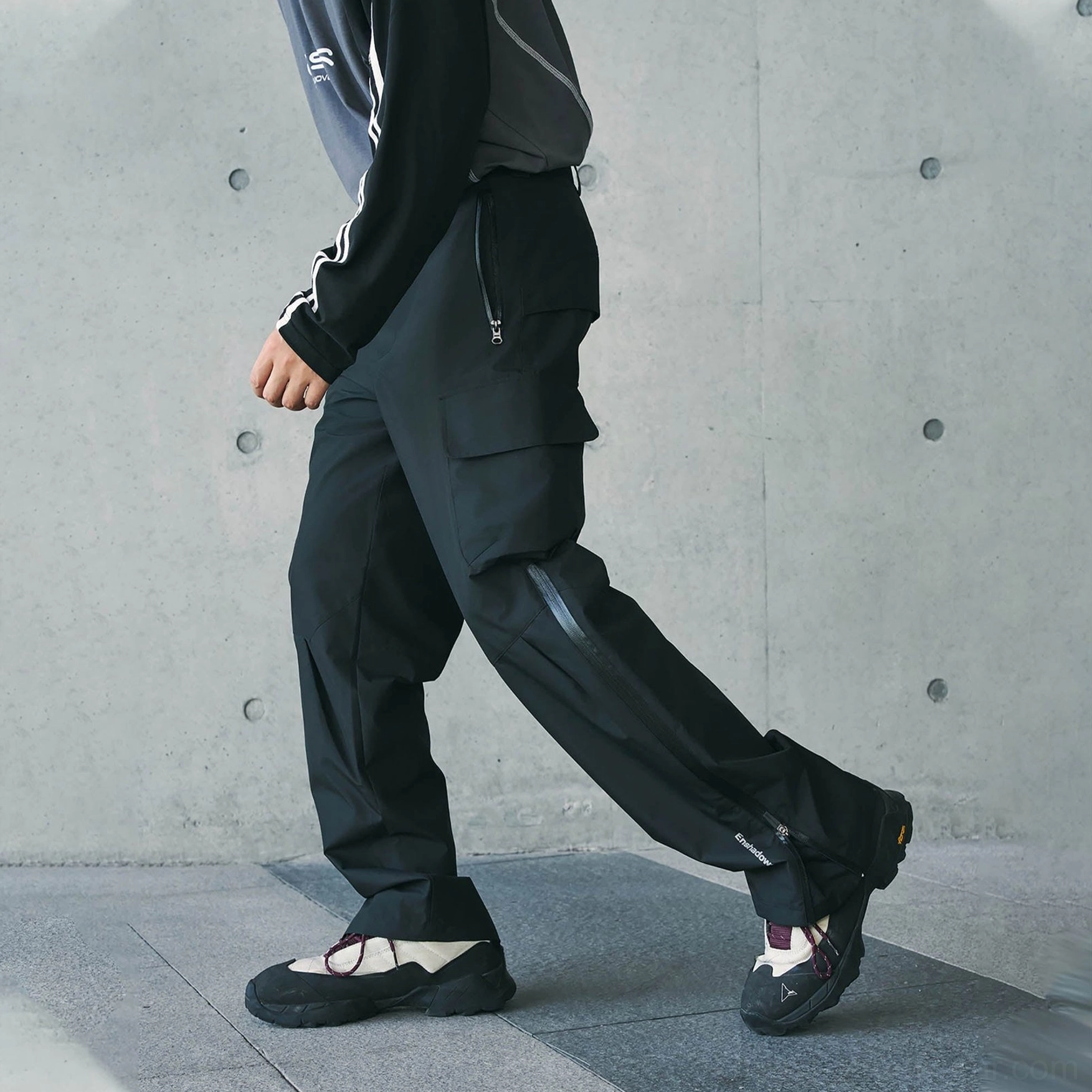 Black techwear cargo pants shown in a dynamic side view with a full-length ankle zipper and oversized cargo pocket
